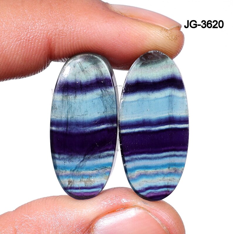 May include: Two oval-shaped gemstones with a layered pattern of blue, purple, and white. The gemstones are held by a person's fingers. The text "JG-3620" is in the upper right corner.
