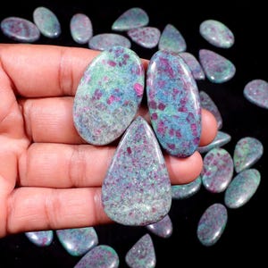 May include: Polished ruby zoisite gemstones in various shapes, including oval and teardrop. The stones display a mix of green, blue, and red colours. The gemstones are displayed against a black background.