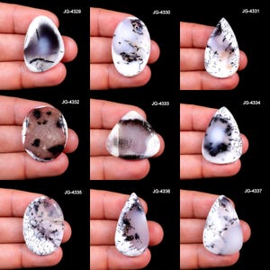 May include: A collection of nine polished gemstones in various shapes, including oval, teardrop, and triangular. The stones feature a white base with intricate black and gray dendritic patterns. Each stone is held in a hand against a black background.