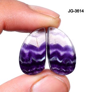 May include: Two polished, teardrop-shaped gemstones with a vibrant purple and white banded pattern. The stones are held between fingers, showcasing their translucent quality. The text "JG-3614" is visible in the upper right corner.