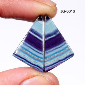 May include: Two triangular-shaped gemstones with a layered pattern of purple, blue, and white. The gemstones are held between fingers and have a polished surface. The text "JG-3616" is visible in the upper right corner.