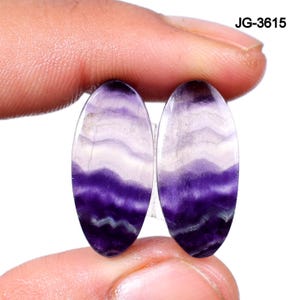 May include: Two oval-shaped gemstones with a layered pattern of purple and white hues. The gemstones are held between fingers, showcasing their polished surfaces and natural color variations. The text "JG-3615" is visible in the upper right corner.