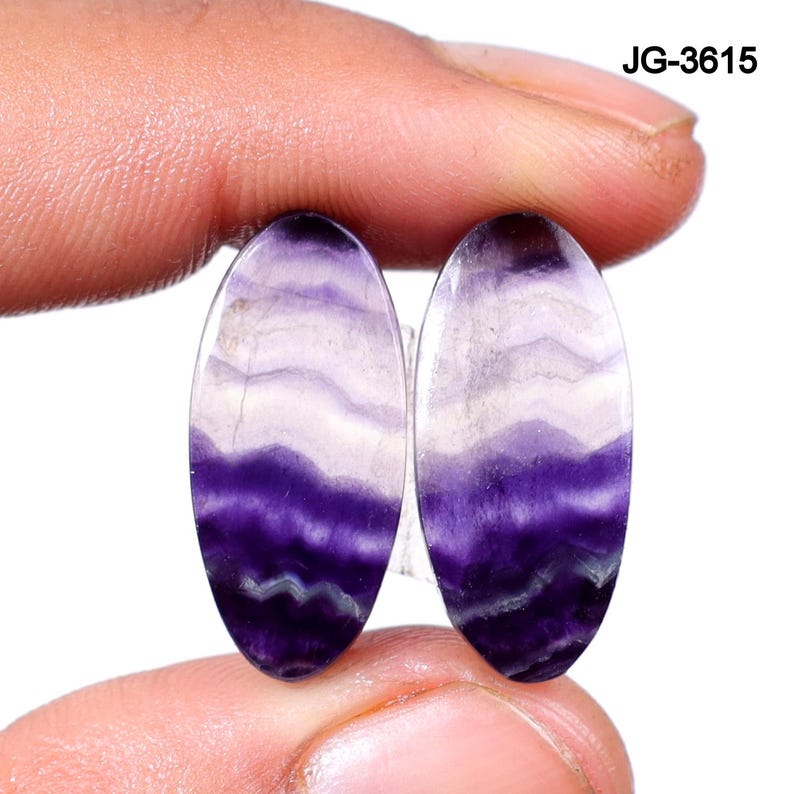 May include: Two oval-shaped gemstones with a layered pattern of purple and white hues. The gemstones are held between fingers, showcasing their polished surfaces and natural color variations. The text "JG-3615" is visible in the upper right corner.