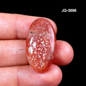 Op de afbeelding: Ovale zonnesteen edelsteen met een sprankelende, rood-oranje tint. De steen heeft een gepolijst oppervlak en vertoont een glittereffect. De tekst "JG-3696" is zichtbaar. De edelsteen wordt in een hand gehouden tegen een zwarte achtergrond.