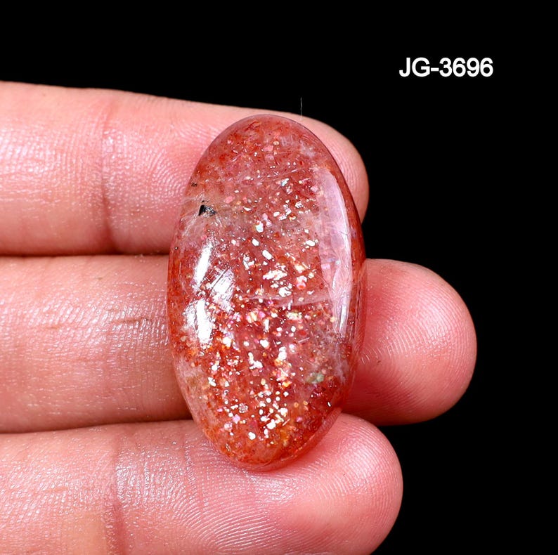 Op de afbeelding: Ovale zonnesteen edelsteen met een sprankelende, rood-oranje tint. De steen heeft een gepolijst oppervlak en vertoont een glittereffect. De tekst "JG-3696" is zichtbaar. De edelsteen wordt in een hand gehouden tegen een zwarte achtergrond.
