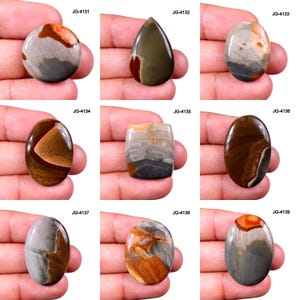 May include: Nine polished gemstone cabochons in various shapes, including round, teardrop, and oval. The stones display a range of colours, such as brown, grey, orange, and cream, with unique patterns and textures. Each stone is labelled with a "JG" code.