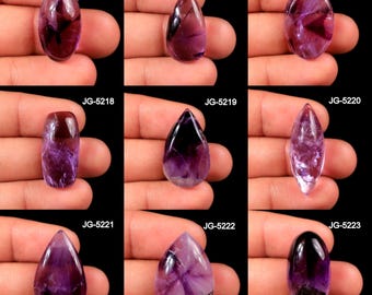 Puede incluir: Nueve piedras preciosas de amatista pulidas en varias formas, incluyendo ovaladas, en forma de lágrima y alargadas. Las piedras muestran una gama de tonos púrpuras, con algunas mostrando inclusiones más oscuras, casi negras. Cada piedra se sostiene en una mano sobre un fondo negro.