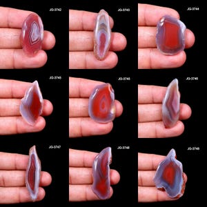 May include: Nine polished agate stones with varying shapes and patterns. The stones display bands of red, white, and gray hues. Each stone is held in a hand against a black background. The product codes JG-3742 through JG-3748 are visible.