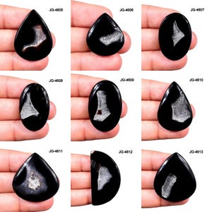May include: A selection of polished black gemstone cabochons, each featuring distinctive white and grey internal patterns. The stones vary in shape, including teardrop, oval, and irregular forms. The gemstones are presented against a neutral backdrop.
