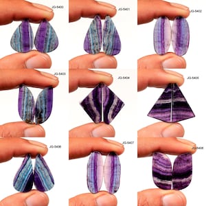 Puede incluir: Surtido de piedras preciosas de fluorita pulida en varias formas, incluyendo lágrima, ovalada y diamante. Las piedras muestran una mezcla de bandas moradas, azules y blancas. Cada par se sostiene entre los dedos, con el código del producto visible.