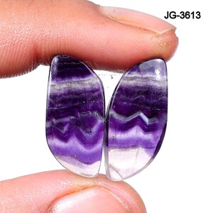 May include: Two polished, semi-precious gemstones with a butterfly wing shape. The stones feature bands of deep purple and white, with a translucent, clear base. The gemstones are held between fingers, and the text "JG-3613" is visible in the upper right corner.