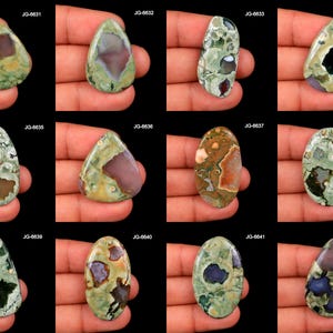 May include: A collection of twelve polished gemstone cabochons in various shapes, including teardrop and oval. The stones display a mix of green, brown, and purple hues, with unique patterns and textures. Each stone is labeled with a "JG" code.