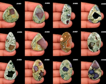 May include: A collection of twelve polished gemstone cabochons in various shapes, including teardrop and oval. The stones display a mix of green, brown, and purple hues, with unique patterns and textures. Each stone is labeled with a "JG" code.