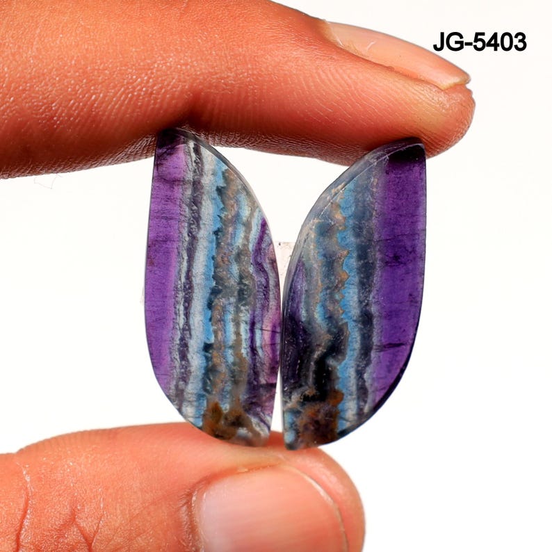 Op de afbeelding: Twee gepolijste edelstenen met een gelaagde uitstraling. De stenen worden tussen de vingers gehouden en vertonen een mix van kleuren, waaronder paars, blauw en bruin. De edelstenen hebben een gladde, gebogen vorm en worden waarschijnlijk gebruikt voor sieraden of decoratieve doeleinden.