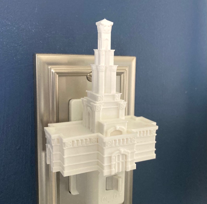 May include: A white, 3D-printed night light shaped like a multi-tiered building with a tall tower. The night light is plugged into a standard wall outlet with an on/off switch. The background is a solid blue wall.