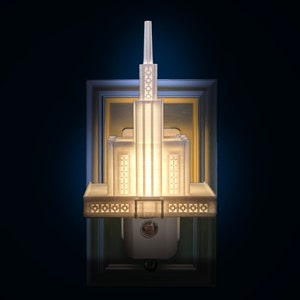 May include: A white, Art Deco-style wall sconce with a rectangular frame. The sconce features a tiered design with geometric patterns and emits a warm, inviting light. The background is a dark blue.