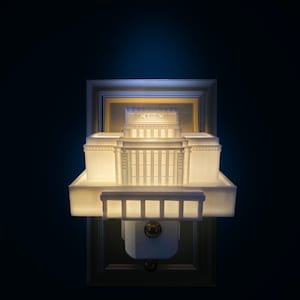 May include: A white architectural model of a building with columns and a tiered roof, illuminated against a dark background. The model is mounted on a rectangular base with a decorative frame, suggesting a wall sconce or night light.