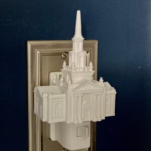 Op de afbeelding: Een wit 3D-geprint nachtlampje in de vorm van een kerkgebouw, met een torenspits en architectonische details. Het nachtlampje is aangesloten op een zilveren stopcontact op een donkerblauwe muur.