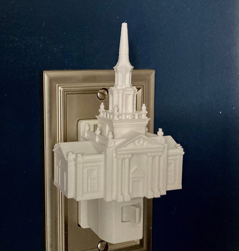 Op de afbeelding: Een wit 3D-geprint nachtlampje in de vorm van een kerkgebouw, met een torenspits en architectonische details. Het nachtlampje is aangesloten op een zilveren stopcontact op een donkerblauwe muur.