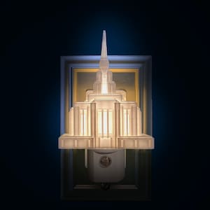 May include: A white architectural night light with a detailed design, resembling a temple or building, is illuminated against a dark blue background. The night light is plugged into a wall outlet. The overall theme is home decor.