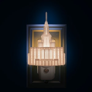 Puede incluir: Una luz de noche blanca con forma de templo, con una alta aguja y múltiples niveles. La luz de noche está montada en una pared y enmarcada en una forma rectangular. La luz está encendida, iluminando los detalles del edificio.