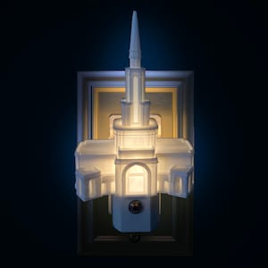 May include: A white architectural night light with a steeple design, mounted on a gold-framed wall plate. The night light is illuminated, casting a warm glow. The design resembles a church or temple.