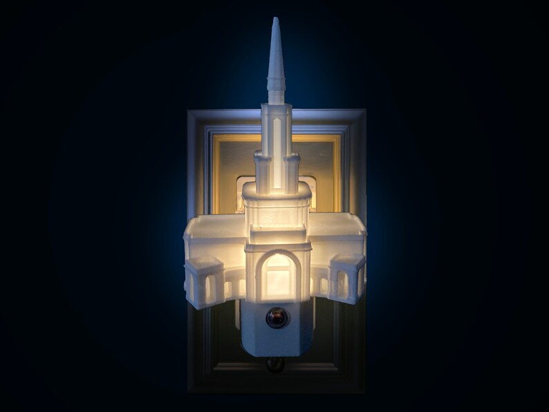 May include: A white architectural night light with a steeple design, mounted on a gold-framed wall plate. The night light is illuminated, casting a warm glow. The design resembles a church or temple.