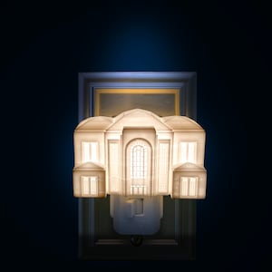 May include: A white, architectural-style night light shaped like a building with illuminated windows and doors. The night light is mounted on a wall outlet and is set against a dark blue background. The design resembles a classical building.