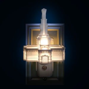 May include: A white architectural night light with a detailed design, resembling a building with a tower. The night light is mounted on a rectangular frame with a gold and white border, and is illuminated from within.