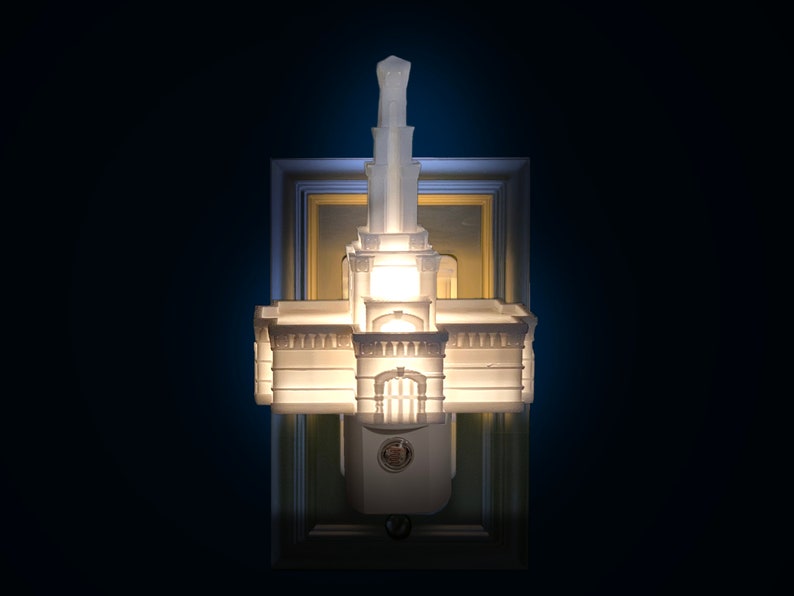 May include: A white architectural night light with a detailed design, resembling a building with a tower. The night light is mounted on a rectangular frame with a gold and white border, and is illuminated from within.