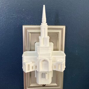 May include: A white architectural night light shaped like a building with a tall spire. The night light is plugged into a wall socket with a silver-coloured plate. The switch on the night light is labelled "ON" and "OFF".