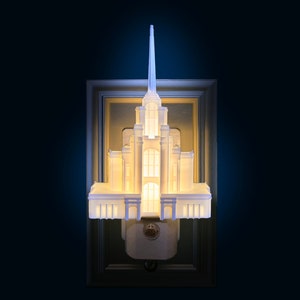 May include: A white architectural night light shaped like a temple, with a tall spire and multiple levels. The night light is mounted on a rectangular frame and emits a soft, warm glow. The background is a dark blue.