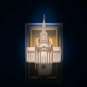Op de afbeelding: Een witte architecturale nachtlamp in de vorm van een gebouw met een torenspits. De nachtlamp is gemonteerd op een stopcontact en omlijst door een decoratieve rand. De afbeelding is verlicht tegen een donkerblauwe achtergrond.