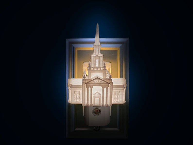 Op de afbeelding: Een witte architecturale nachtlamp in de vorm van een gebouw met een torenspits. De nachtlamp is gemonteerd op een stopcontact en omlijst door een decoratieve rand. De afbeelding is verlicht tegen een donkerblauwe achtergrond.