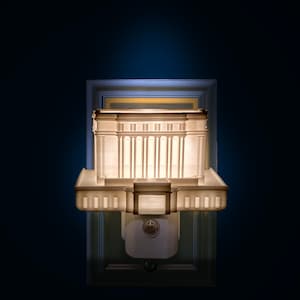 May include: A detailed, illuminated architectural model, possibly a temple or building, with a classical design. The model is white and features columns and a rectangular shape. It is mounted on a wall with a dark blue background.
