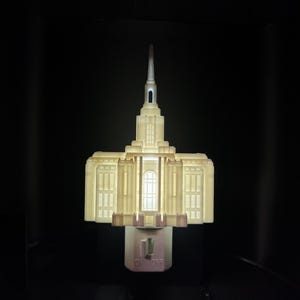 Puede incluir: Una luz de noche de color crema con forma de templo, con una alta aguja y múltiples ventanas. La luz de noche tiene un interruptor de encendido/apagado y está iluminada desde adentro, sobre un fondo oscuro.