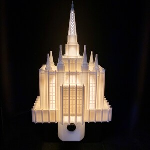 May include: A white, illuminated architectural model of a temple, featuring multiple spires and a rectangular base. The model is lit from within, creating a glowing effect against a black background. The design includes detailed windows and architectural elements.