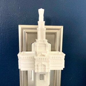 May include: A white architectural night light featuring a detailed building design. The night light has an "ON OFF" switch and is plugged into a wall outlet. The design includes columns, arches, and a tall spire.