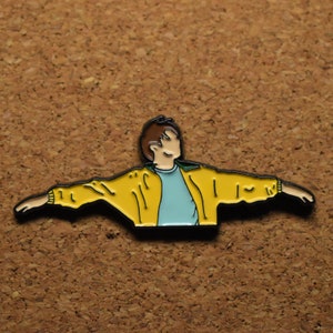 May include: A yellow enamel pin of a person with brown hair wearing a yellow jacket and a light blue shirt with their arms outstretched.