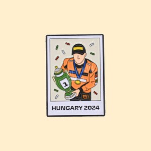 Può includere: Uno spillo in smalto bianco e nero con un'illustrazione di cartone animato di una persona che tiene un trofeo. La persona indossa una tuta da corsa e una medaglia d'oro. Lo spillo ha il testo "HUNGARY 2024" in basso.