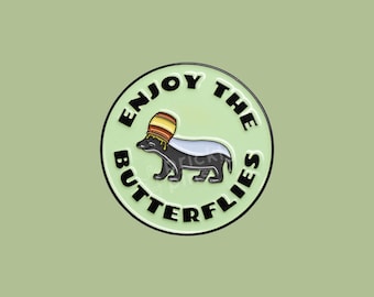 May include: A round enamel pin with a black and white badger wearing a honey pot on its head. The pin says "Enjoy the Butterflies" in black text around the edge.