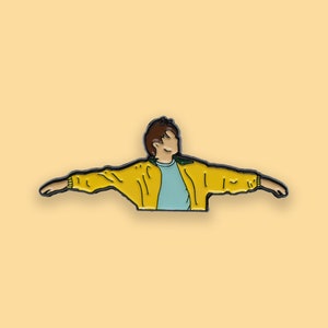 May include: A yellow enamel pin of a cartoon person with brown hair wearing a yellow jacket with green accents and a light blue shirt, with arms outstretched.