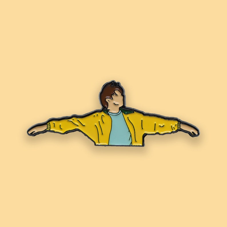 May include: A yellow enamel pin of a cartoon person with brown hair wearing a yellow jacket with green accents and a light blue shirt, with arms outstretched.