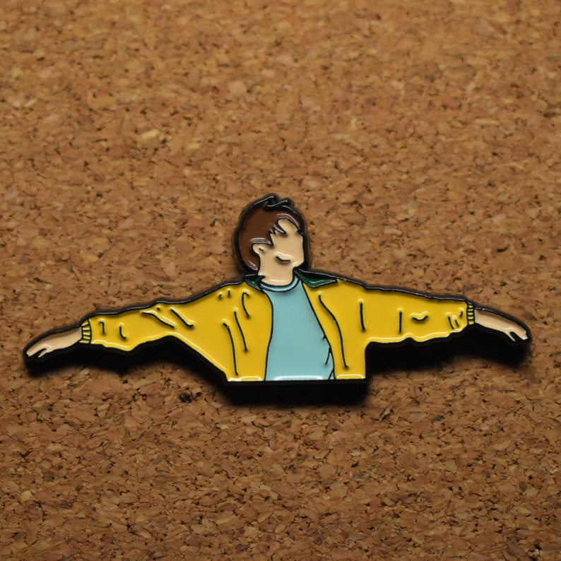 May include: A yellow enamel pin of a person with brown hair wearing a yellow jacket and a light blue shirt with their arms outstretched.