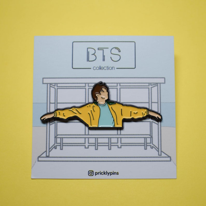 May include: A yellow and blue enamel pin featuring a cartoon figure with arms outstretched, standing in a bus stop. The pin is part of a BTS collection.
