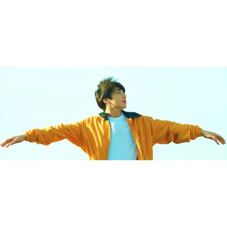 May include: A young man with dark hair wearing a yellow jacket with black trim, standing with arms outstretched against a light blue sky.