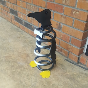 May include: A black metal penguin sculpture with a white bow tie, made from a coiled spring. The penguin has yellow feet and is standing on a concrete surface.