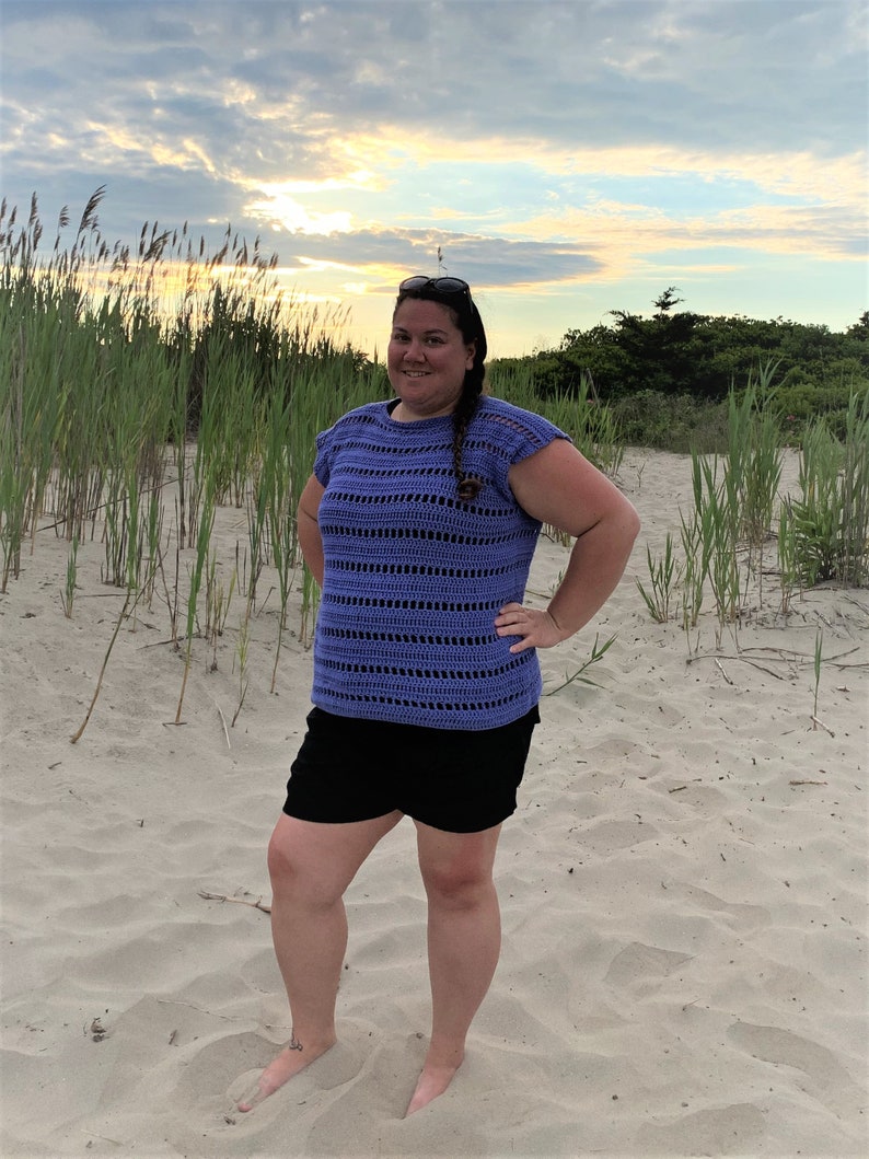 Puede incluir: Una mujer con un top de crochet morado con ribete de crochet negro y pantalones cortos negros. Est&aacute; de pie en una playa de arena con hierba alta en el fondo.