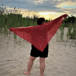 May include: A person wearing a red crocheted shawl with a textured pattern, standing on a sandy beach with tall grass in the background.