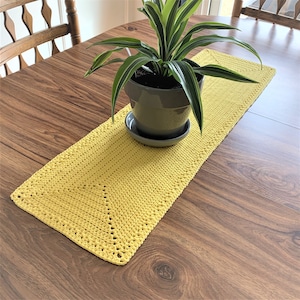 Puede incluir: Un camino de mesa de crochet amarillo con una planta en una maceta gris sobre una mesa de madera.